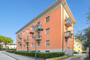 Un edificio residencial de varios pisos con balcones, una calle al frente y un cielo azul despejado.
