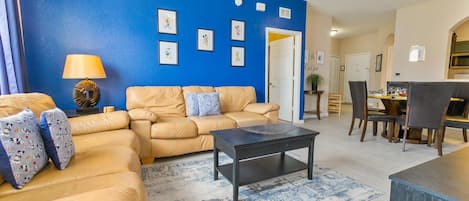 A living room with a blue accent wall, a beige sofa, a coffee table, and a dining area with chairs and a table.