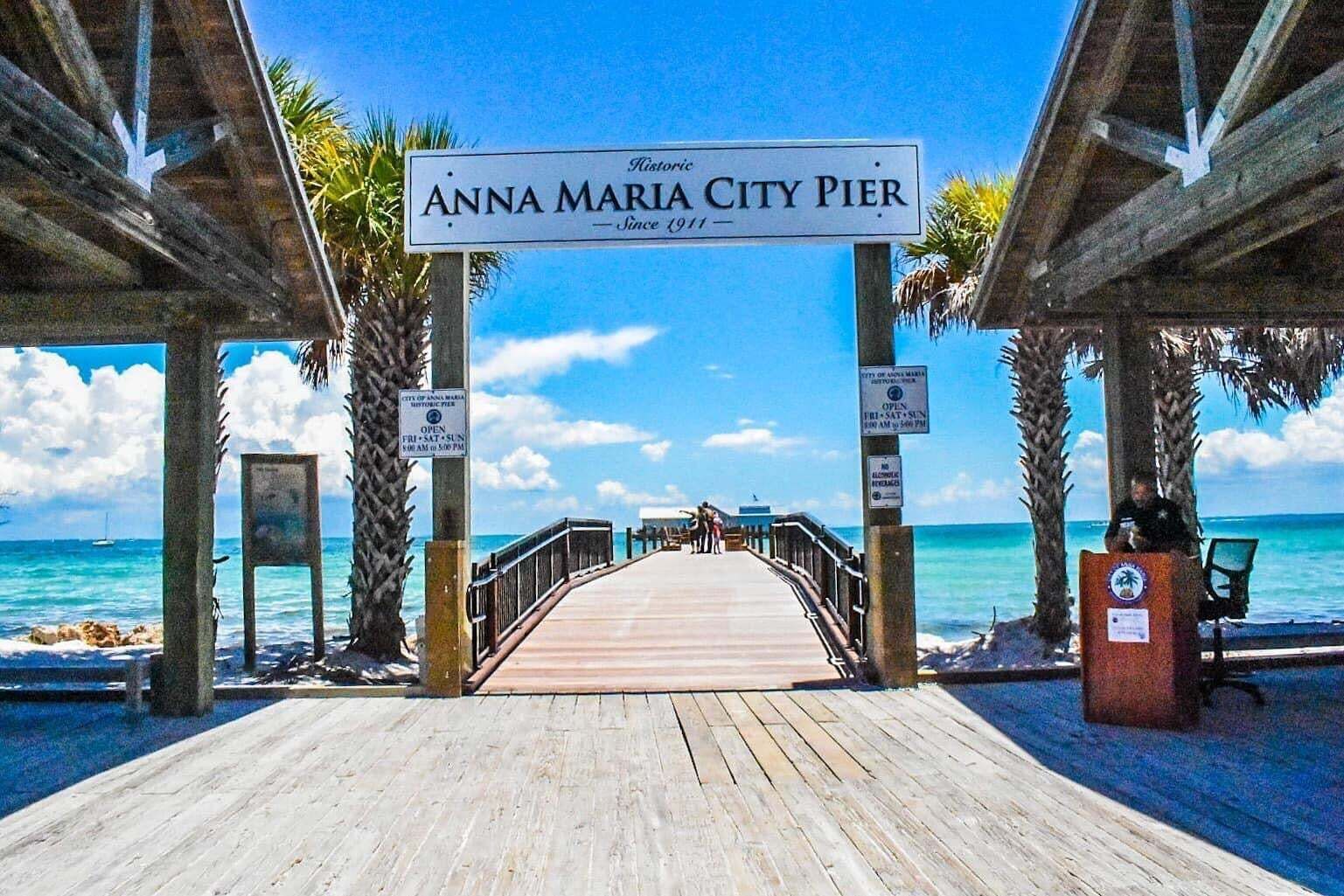 Un muelle de madera que conduce al muelle de la ciudad de Anna Maria, con palmeras y el océano al fondo.