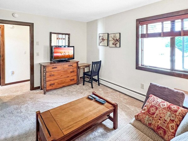 Una sala de estar con una mesa de centro de madera, un televisor sobre un aparador, una silla y una ventana con persianas.