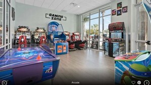 Un salón de juegos interior con diversas máquinas recreativas, incluyendo mesas de hockey de aire y máquinas de pinzas, en una sala amplia con ventanas grandes.