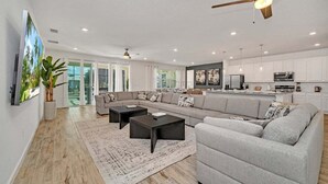 A spacious living area with a large sofa, two coffee tables, and a television. There is a kitchen area with modern appliances and a ceiling fan. - 2525 Shanti Amazing 15 Suites Mansion (Kissimmee)