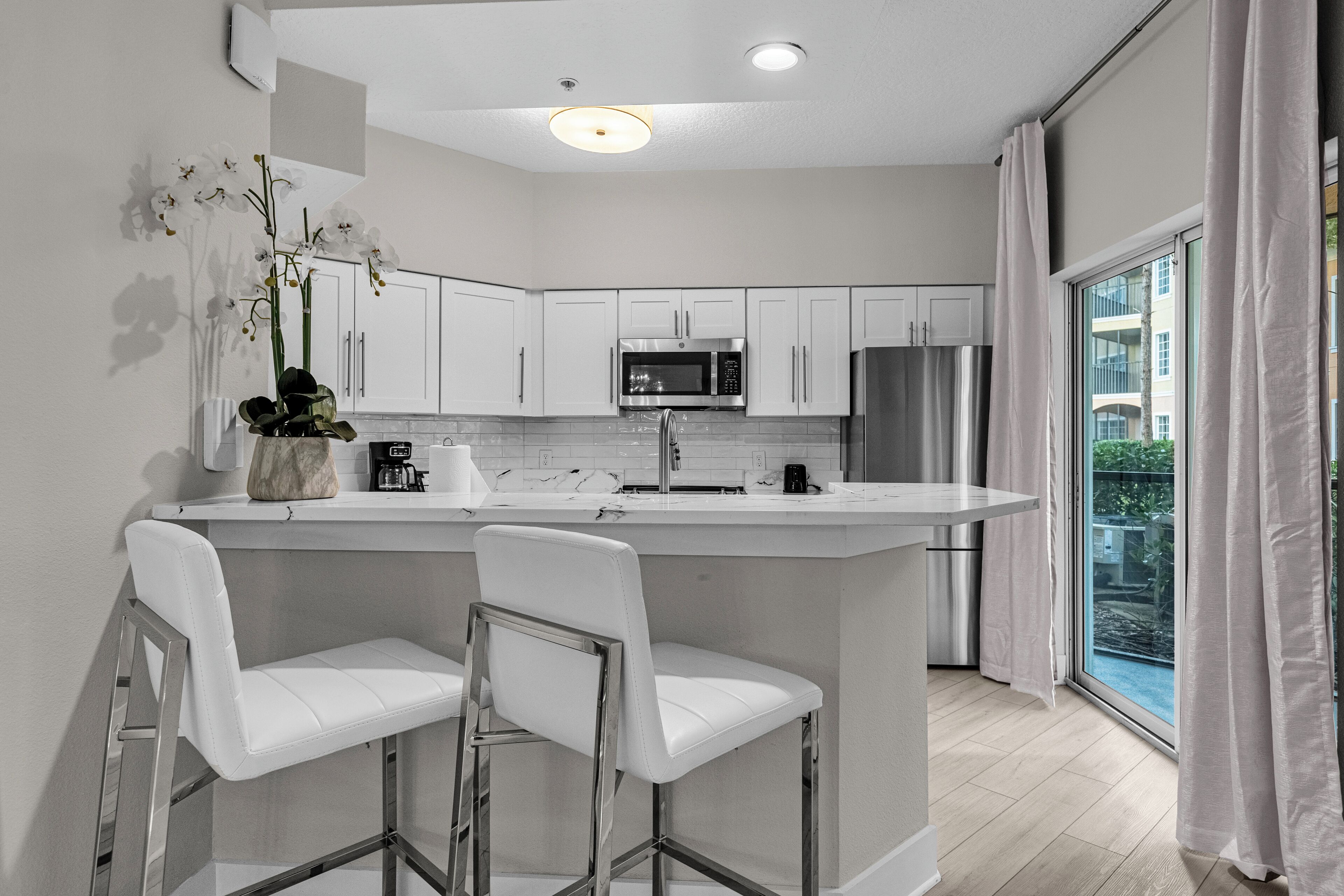 A modern kitchen with white cabinetry, stainless steel appliances, a central island with bar stools, and a large window with curtains.