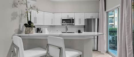 A modern kitchen with white cabinetry, stainless steel appliances, a central island with bar stools, and a large window with curtains.