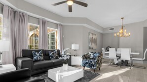A modern living room with a black sofa, white coffee table, and patterned armchair. There is a dining area with white chairs and a chandelier. - 7108 (Orlando)