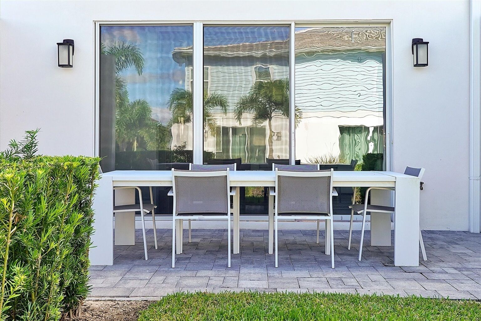 Un espacio comedor exterior blanco con una mesa y cuatro sillas, ubicado frente a un ventanal con vistas a palmeras y un edificio.