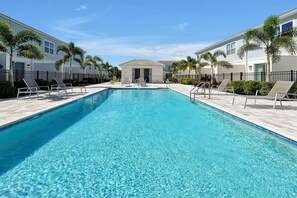A rectangular swimming pool with clear blue water, surrounded by lounge chairs and palm trees, with buildings in the background. - 7909 Draw Street 3bed (Kissimmee)