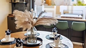 Una mesa de comedor con un jarrón de plumas de pampa, un jarrón de vidrio y una taza. Hay sillas alrededor de la mesa, y se ve un área de cocina al fondo.