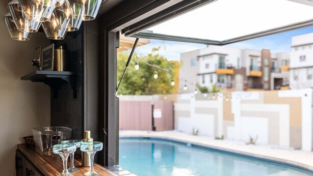 Un bar junto a la piscina con vasos y una botella, vista a una alberca y edificios modernos al fondo.