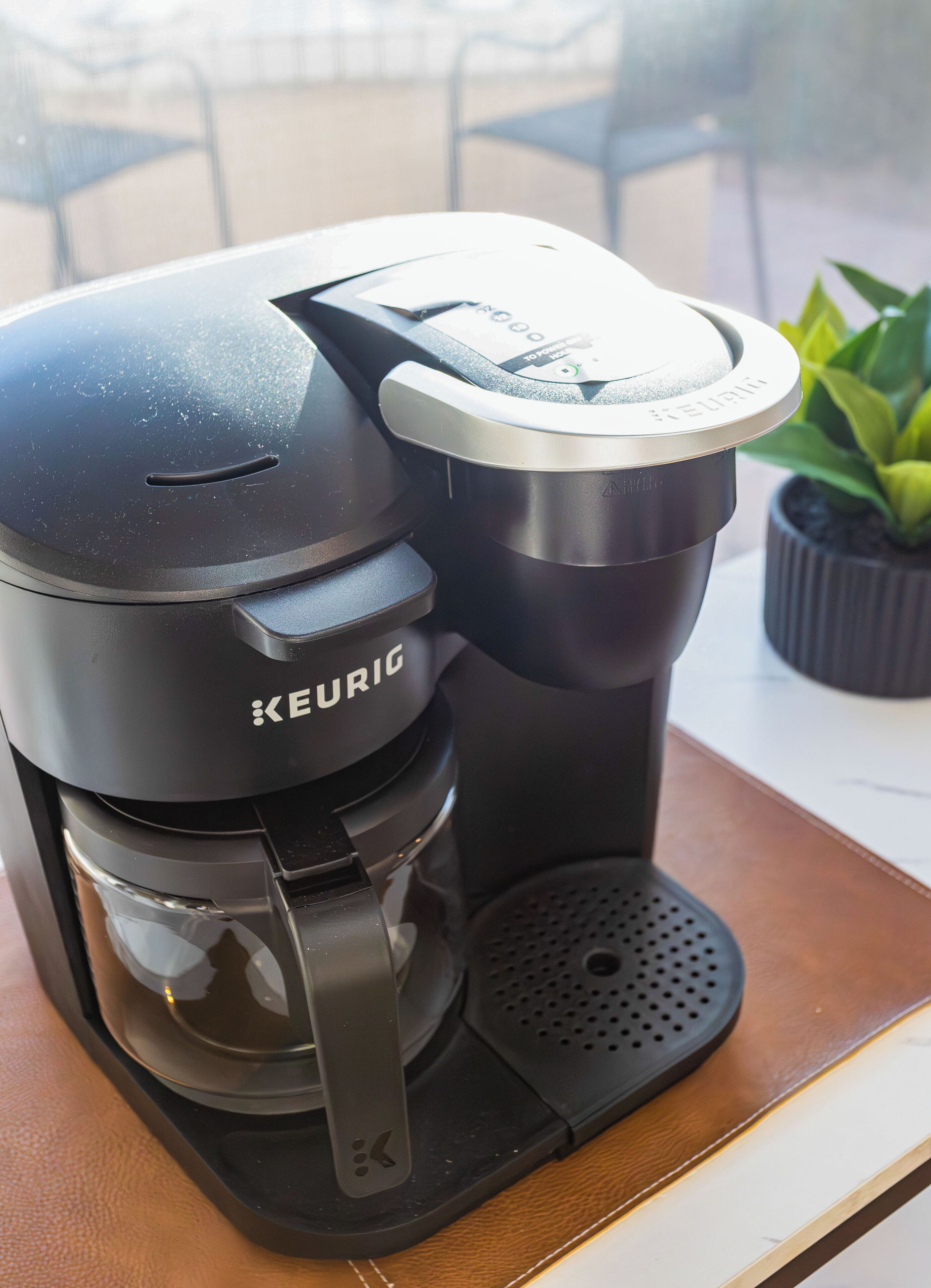 Une machine à café Keurig est posée sur une table, avec une plante en pot en arrière-plan.