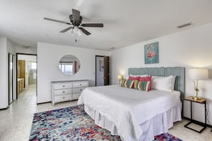 Ein Schlafzimmer mit Bett, Deckenventilator, Kommode, Spiegel, farbenfrohem Teppich und einem Gemälde an der Wand.