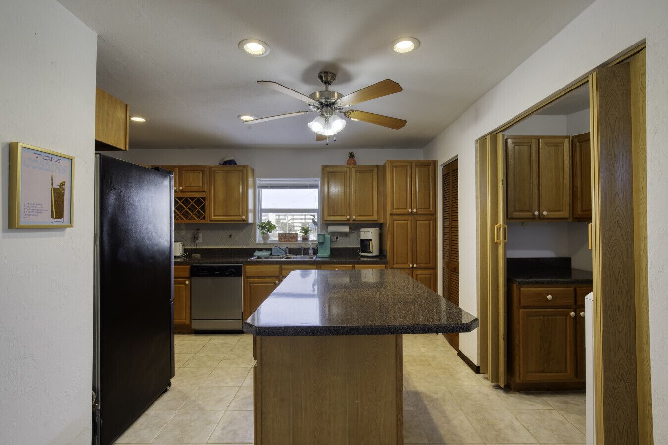 A kitchen with wooden cabinets, a granite countertop, a black refrigerator, a dishwasher, and a ceiling fan.