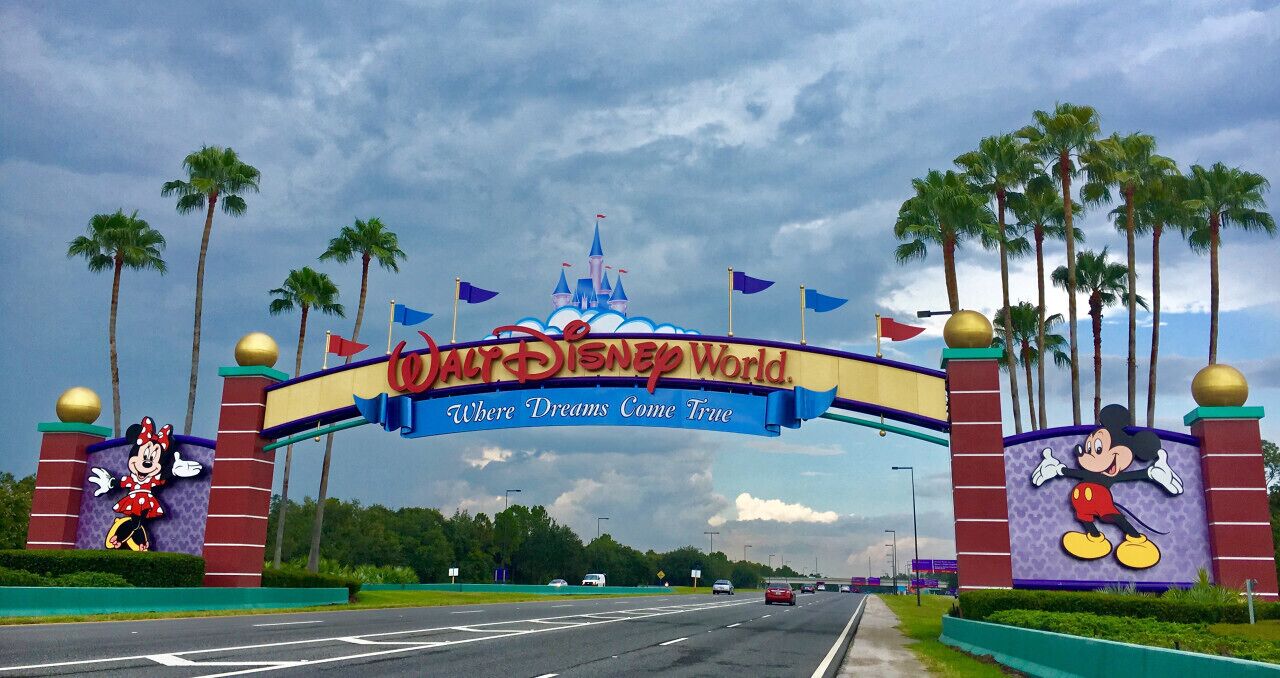 La entrada a Walt Disney World con un gran letrero que muestra a Mickey Mouse y Minnie Mouse, palmeras y un castillo al fondo.