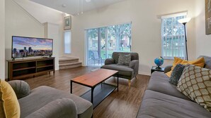 A living room with a flat-screen TV, a sofa, a coffee table, and a staircase leading to another level. - 8460 Crystal Cove Loop 3bed (Kissimmee)