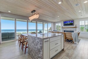 A modern kitchen with a large island, wooden ceiling, and large windows overlooking the ocean.