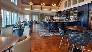 Een ruime barruimte met hoge plafonds en grote ramen, een zithoek met sofa’s en stoelen, en een bar met flessenkasten.