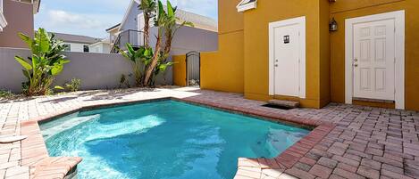 A swimming pool with a brick border, surrounded by a yellow building with white doors and a small gate.
