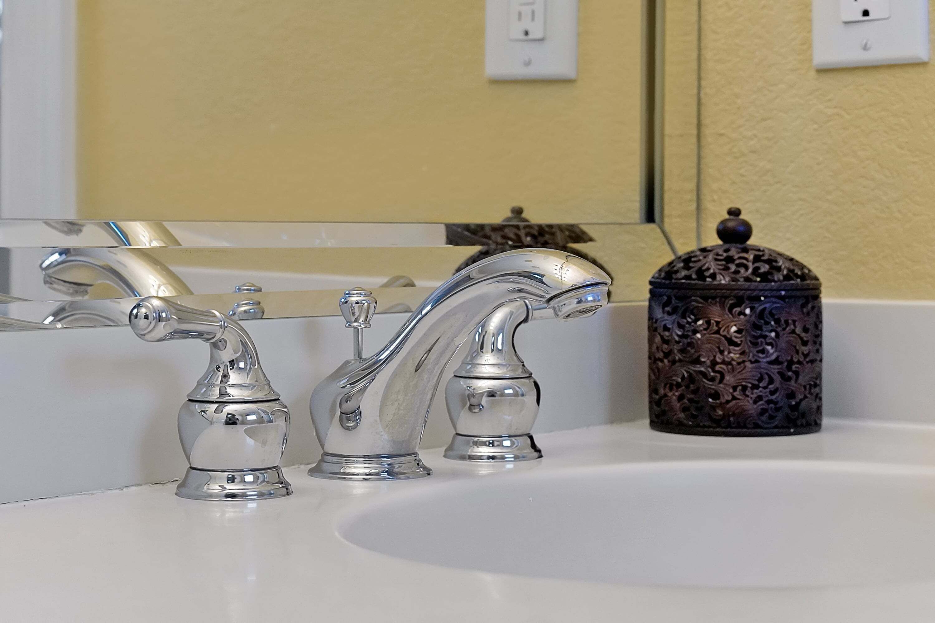 A bathroom with a white sink, two chrome faucets, and a decorative container.