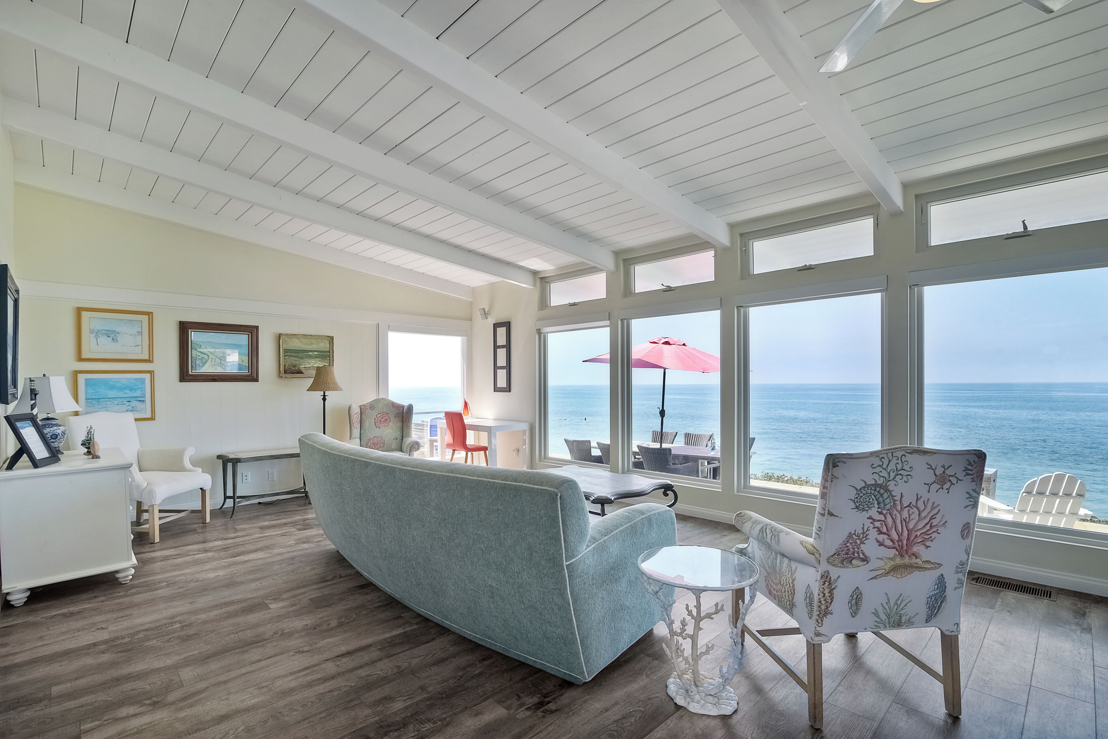 Un salón amplio con un gran ventanal con vistas al océano, un sofá azul y una silla blanca con estampado coral.