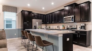 A modern kitchen with dark cabinetry, a central island with bar stools, a refrigerator, microwave, stove, and a coffee maker. - Adt8972 (Kissimmee)