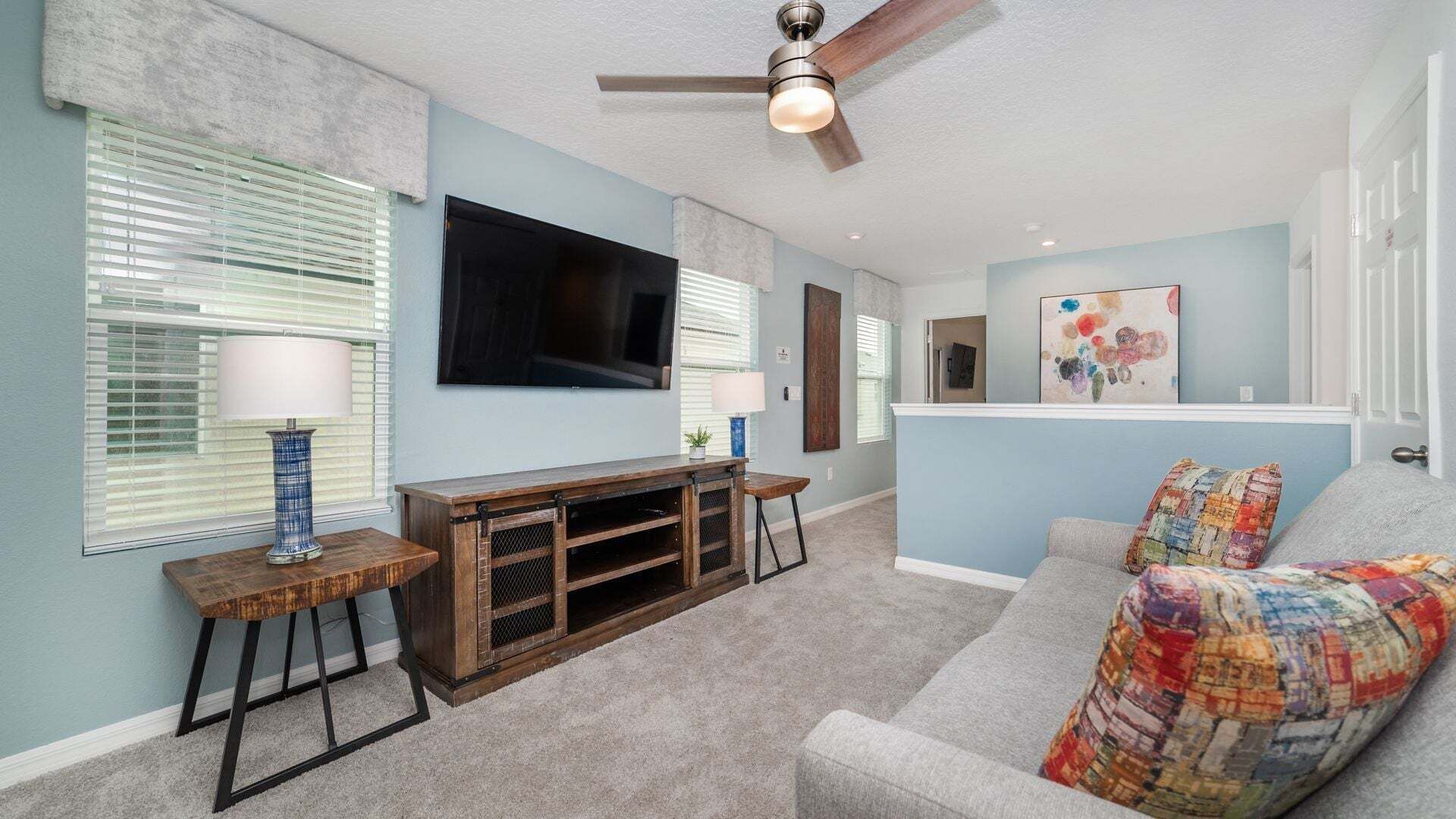 A living room with a ceiling fan, a flat-screen TV, a wooden entertainment unit, a sofa with a colorful throw, and a painting on the wall.