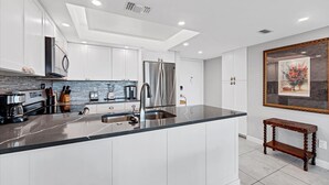 A modern kitchen with white cabinetry, a stainless steel refrigerator, a marble countertop, and a small wooden bench. - Anchorage Resort 8th Floor Condo in Siesta Key With Private Beach Access, Pool, Boat Dock 0894 (Siesta Key)