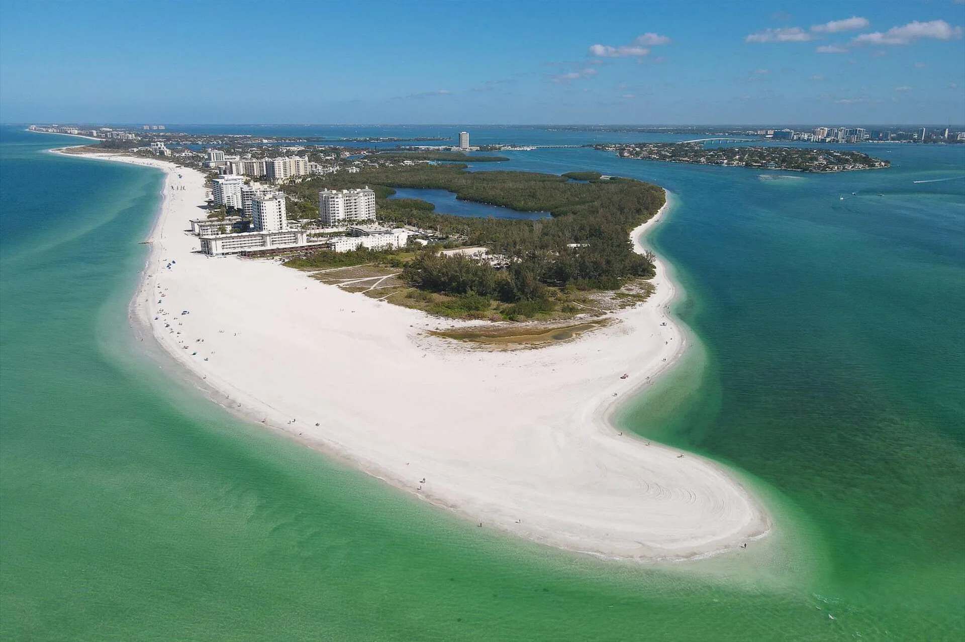 An aerial view of a coastal area with a long sandy beach, a cluster of high-rise buildings, and a small island with dense vegetation.