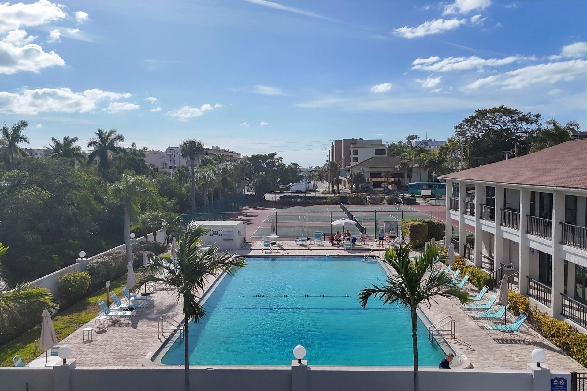 A swimming pool with lounge chairs and palm trees, surrounded by buildings and a clear sky.