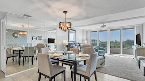 A modern dining area with a glass table, chairs, and a chandelier. There is a living area with a sofa and a TV. The room has large windows and a ceiling fan. - Anchorage Resort Siesta Key 6th Floor 2 Bedroom Condo Beach Access 0890 (Siesta Key)