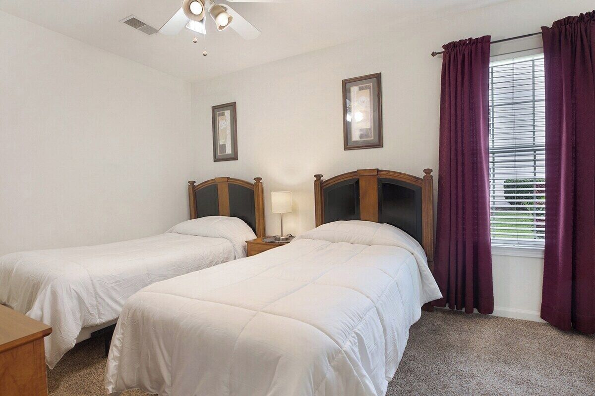 A bedroom with two single beds, a nightstand with a lamp, a ceiling fan, and a window with curtains.