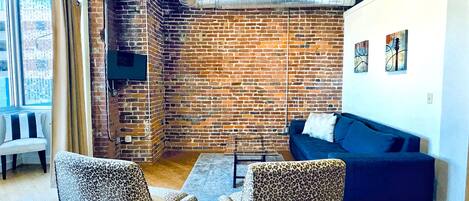 A modern living room with a brick wall, a blue sofa, leopard print chairs, and a flat-screen TV mounted on the wall.