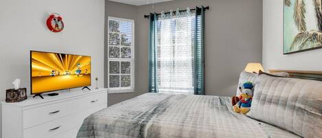 A bedroom with a bed, a TV on a dresser, a Mickey Mouse alarm clock, and a window with curtains.