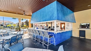 Un área de bar al aire libre con mostrador azul, asientos con sillas de metal y vistas de los edificios circundantes.