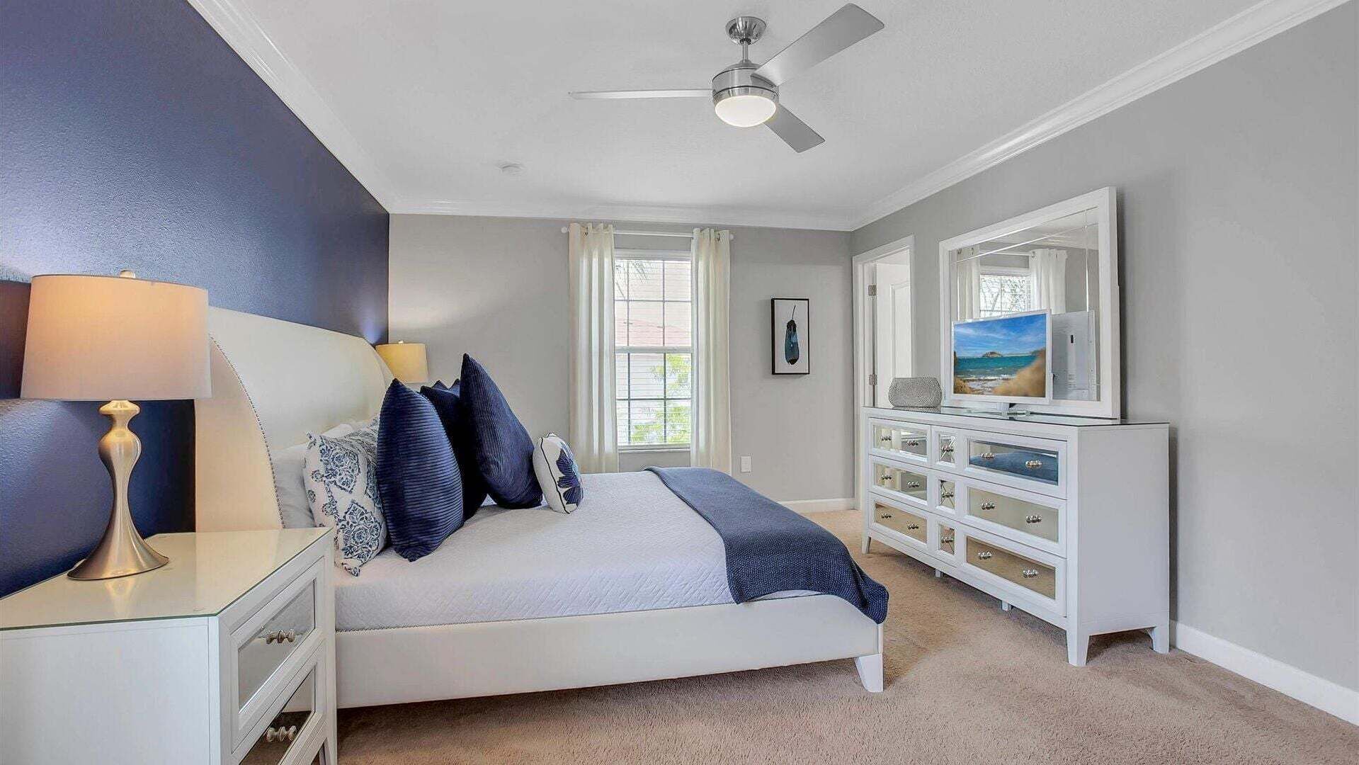 Une chambre à coucher comprenant un lit, une table de chevet, une commode, un ventilateur de plafond et une grande fenêtre.