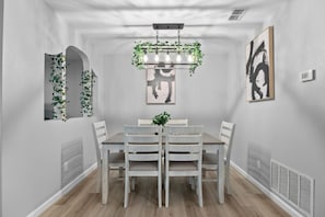 Ein Esszimmer mit weißem Tisch, weißen Stühlen, einer Kronleuchter mit hängenden Pflanzen und einem gerahmten abstrakten Gemälde an der Wand.