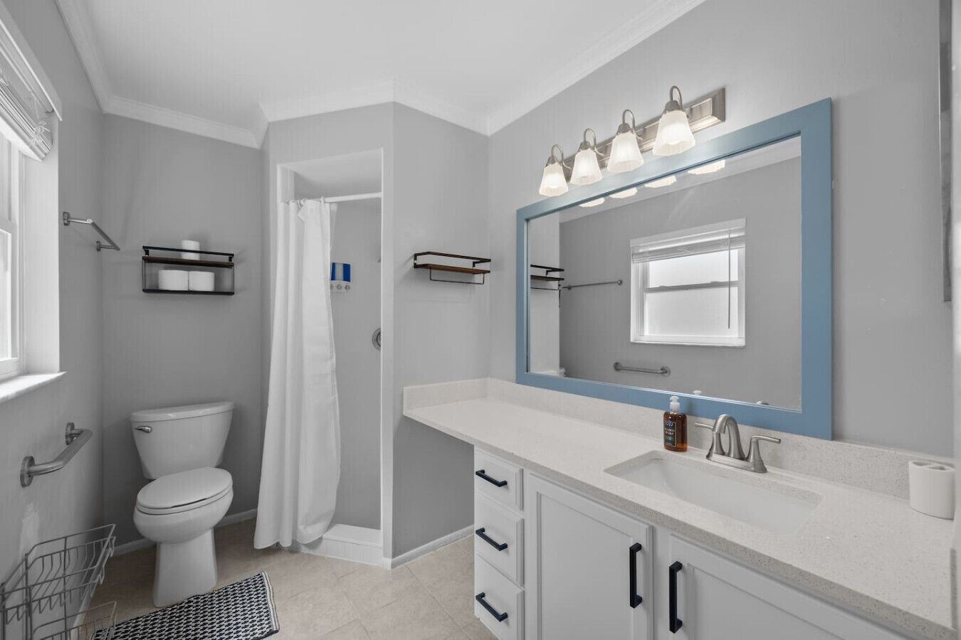 A modern bathroom with a white toilet, a shower with a white curtain, a large mirror with lights, and a white sink with a soap dispenser.