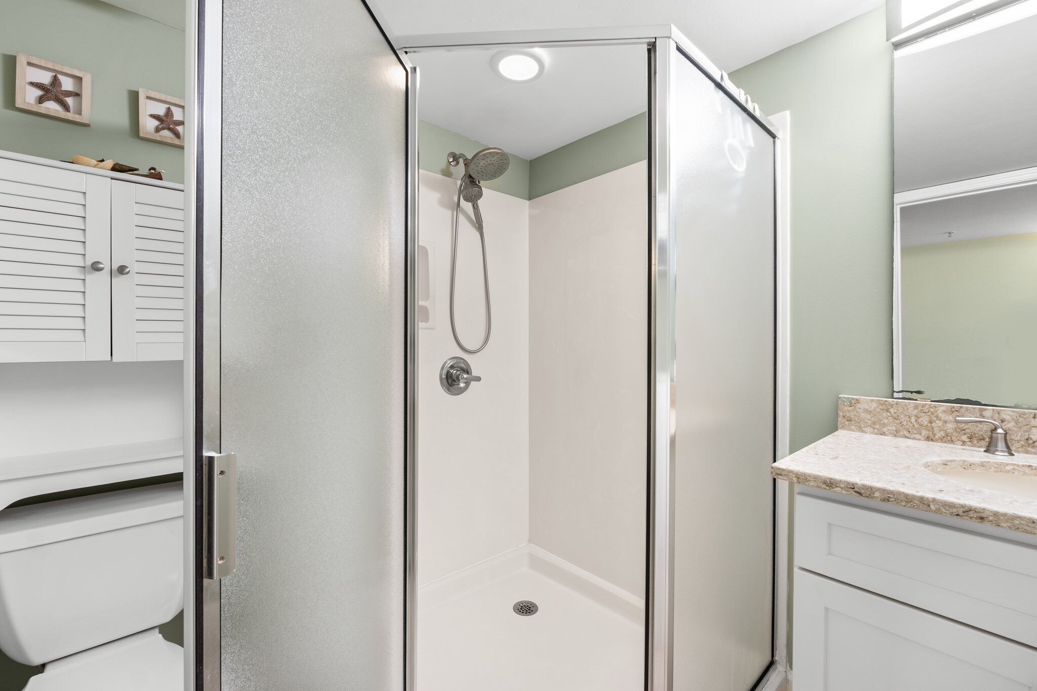 A bathroom with a glass-enclosed shower, a sink with a marble countertop, and a white cabinet with starfish decorations.