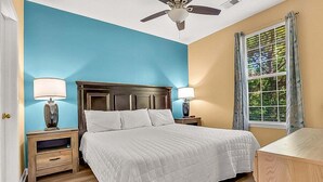 A bedroom with a large bed, wooden headboard, two bedside tables with lamps, a window with curtains, and a ceiling fan. - River Oaks 60N (Myrtle Beach)