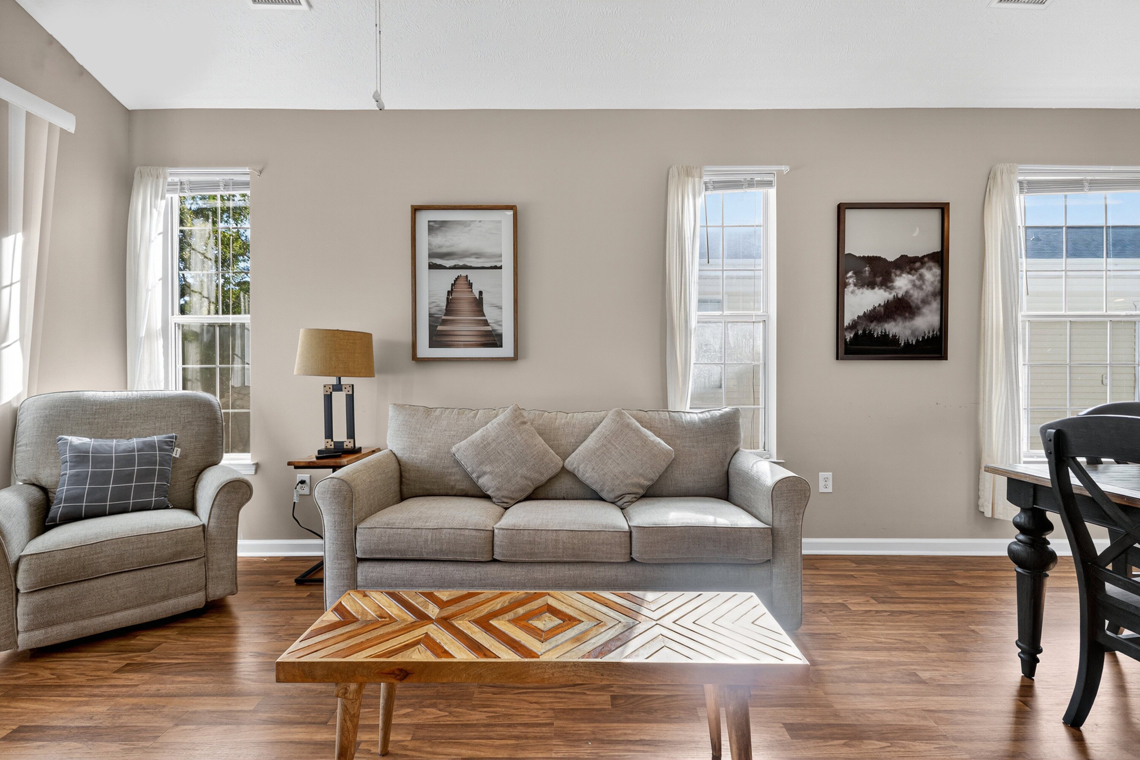 A living room with a sofa, armchair, and coffee table, featuring artwork on the walls and a lamp.