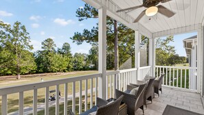 A covered porch with wicker furniture and a ceiling fan, overlooking a grassy area with trees and a road. - River Oaks 52I (Myrtle Beach)