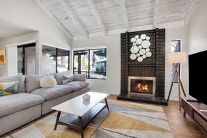 A modern living room with a stone fireplace, a grey sofa, a white coffee table, and a TV on a wooden stand. - Pueblo Vista (Palm Springs)
