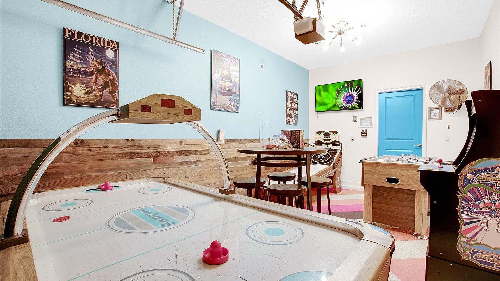 A room with a shuffleboard table, a foosball table, and a dartboard. There are posters on the walls and a blue door.