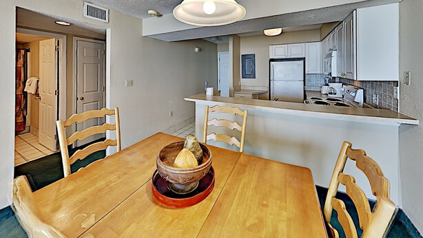 A dining area with a wooden table and chairs, a bowl of fruit, and a kitchen in the background. - Sands Beach Club 123 (Myrtle Beach)