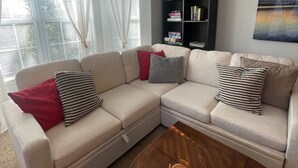 A living room with a beige sectional sofa, a coffee table, a bookshelf, and a painting on the wall. - Putters Hideaway @ Legends Golf Resort (Myrtle Beach)