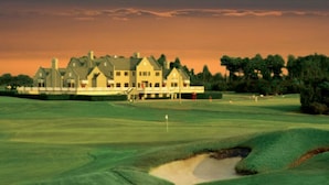 A large, multi-story building with a symmetrical facade, likely a clubhouse or hotel, situated on a golf course with a well-maintained green and sand bunkers. - Putters Hideaway @ Legends Golf Resort (Myrtle Beach)