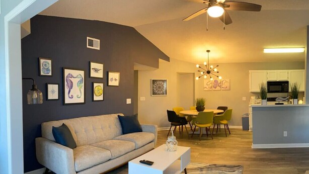 A modern living room with a sofa, coffee table, and artwork on the wall. There is a dining area with chairs and a chandelier.