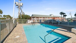 Ein Schwimmbecken mit weißem Zaun, ein Gebäude mit grüner Dachbegrünung und Palmen im Hintergrund.