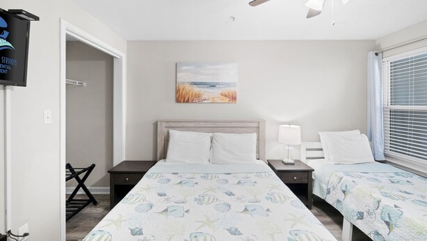 Ein Schlafzimmer mit zwei Betten, einem Deckenventilator, einem Strandgemälde und einem Fenster mit Jalousien.