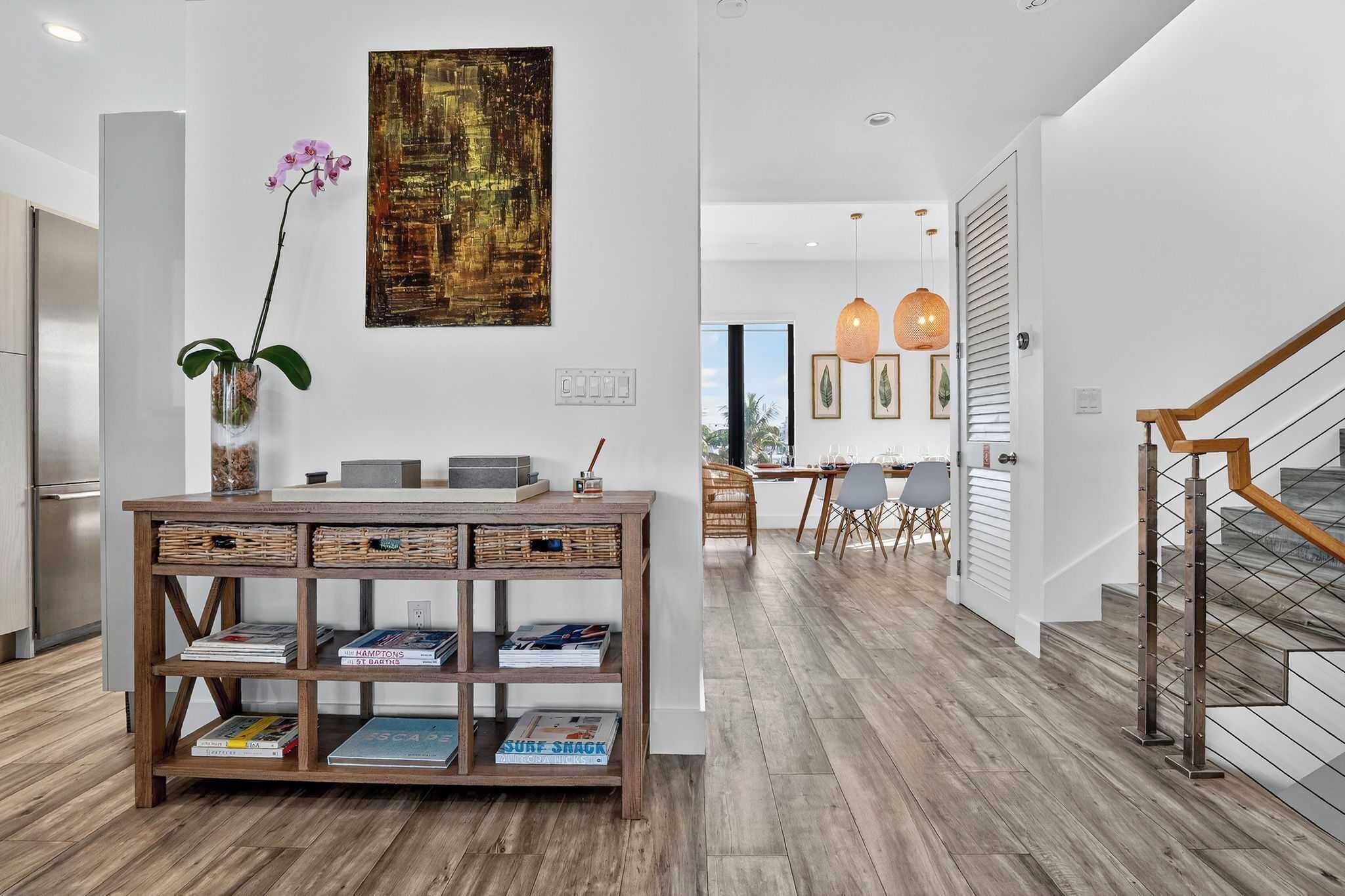 A modern kitchen with wooden flooring, a wooden console table with books, a vase with flowers, and a large abstract painting on the wall.