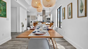 A modern dining area with a wooden table set for six, white walls, and a kitchen in the background. - Shoreline Villa by Rove Rooftop & Beach Access (Hollywood)